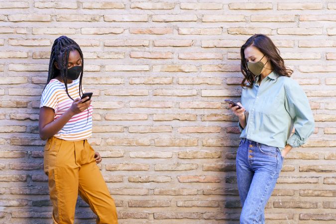 Two girls with facemasks standing apart holding phone beside wall
