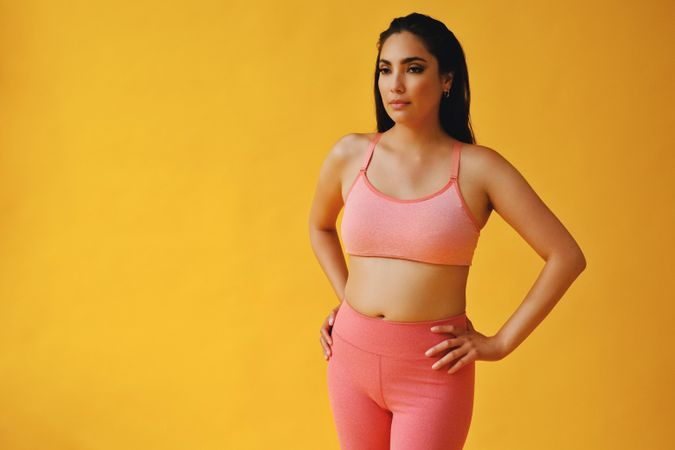Hispanic woman in yoga clothes standing in yellow studio with hands on hip, copy space