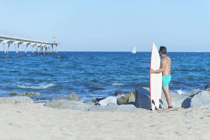 Back of male surfer standing on rock near sea with board