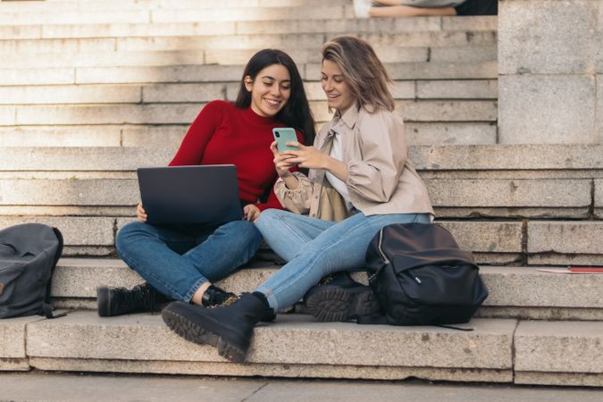 Two students sitting on the stairs of the college with the laptop and the mobile