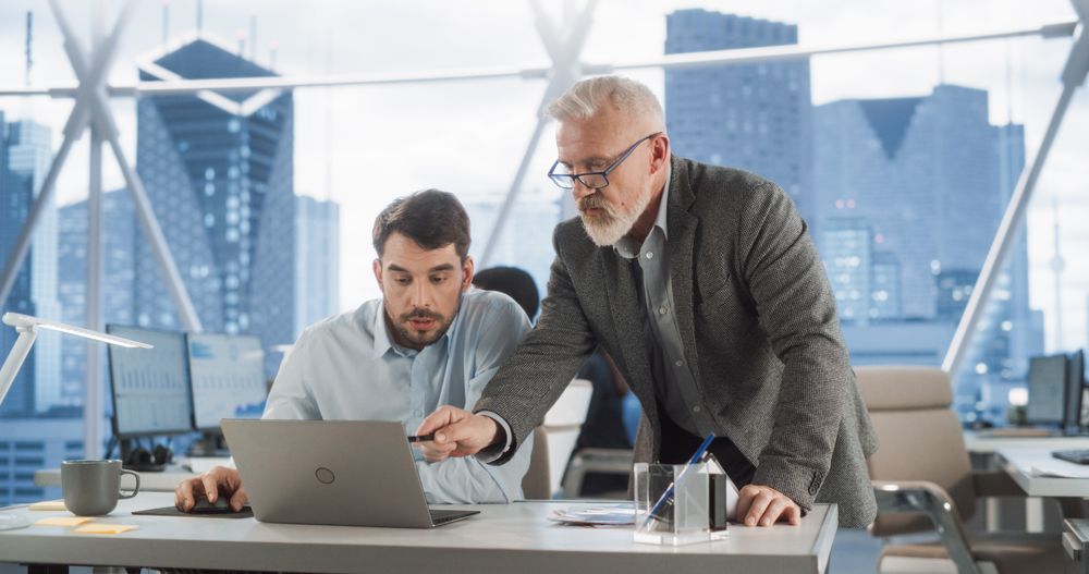 Male CEO Talking to Young Financial Strategist Behind Laptop In Skyscraper Office In Downtown.