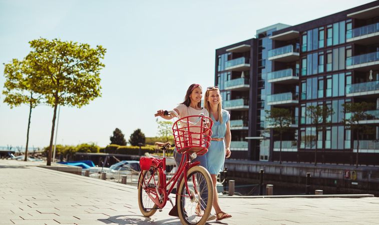 Two close female friends happily walking along the riverwalk with red bike