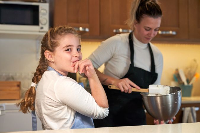 Girl in apron licking her hand in the kitchen with a girl in the background mixing ingredients in a metal container