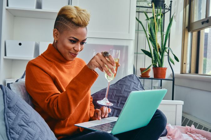 Happy Black woman smiling at a laptop toasting with champagne in a bright modern loft