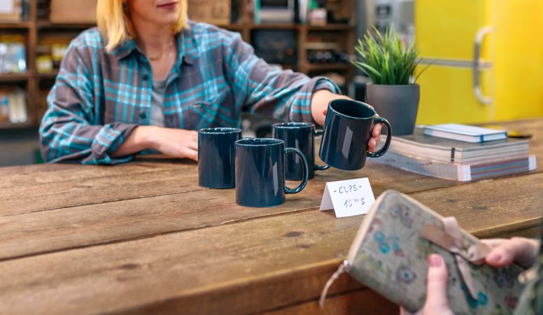 unrecognizable woman employee with plaid shirt showing blue mugs to customer in vintage shop