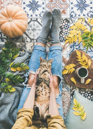 Woman with cat on lap sitting on colorfully tiled balcony with fall leaves, squash, and mug