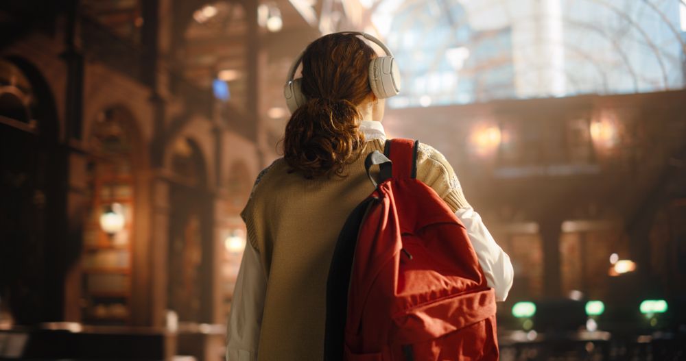 Young Female Student With Headphones and Red Backpack Walking in a Grand Library