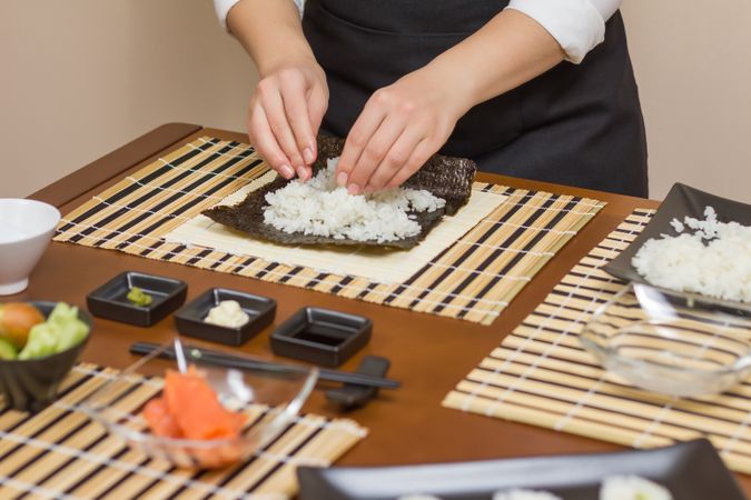 Chef in apron filling Japanese sushi rolls with rice in a nori sheet on a traditional mat