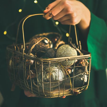 Woman in green dress holding wire basket of gold holiday decorations, selective focus, square crop