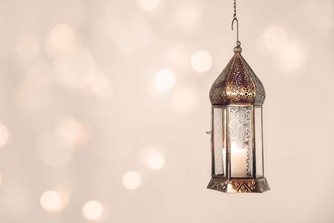 Hanging copper burning Moroccan lantern. Festive pale beige background with glittering bokeh lights. Ramadan Kareem muslim holiday still life. Neutral blurred background. Elegant Iftar dinner.