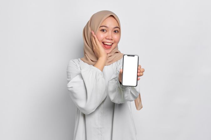 Asian Muslim woman in a bright studio shoot holding cell phone and smiling with hand on cheek