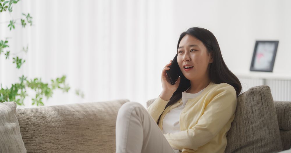 Sitting on the Sofa in Her Cozy Apartment, Beautiful Smiling South Korean Woman is Having a Thoughtful Phone Conversation