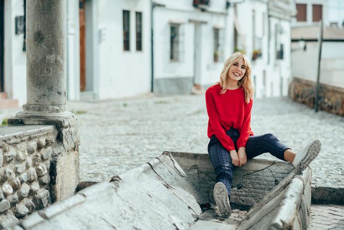 Happy young blond woman sitting on cement trough in Spanish town