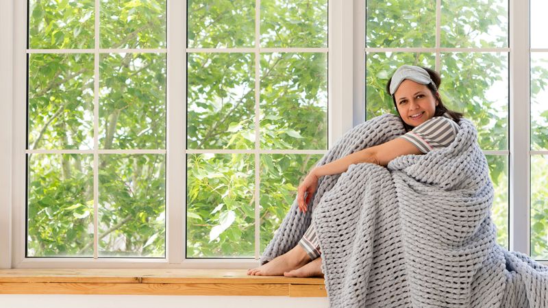 Woman sitting by window, enveloped in cozy gray blanket.