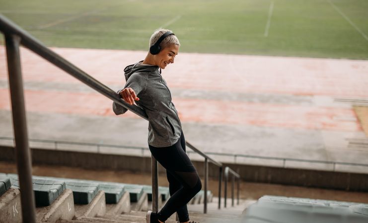 Smiling woman in fitness clothes standing on the stairs of a stadium listening to music