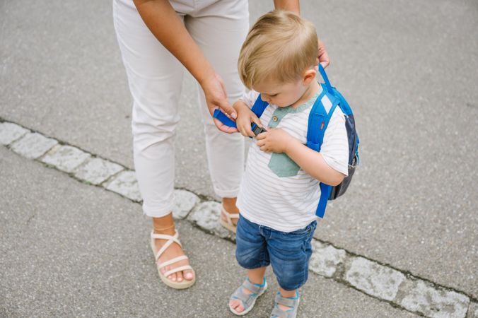 Unrecognizable mother helping child with backpack while standing on asphalt road in daylight