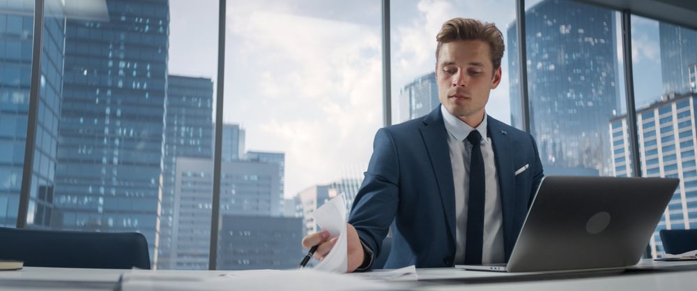 Confident Businessman in a Suit Sitting at a Desk in Modern Office, Using Laptop Computer