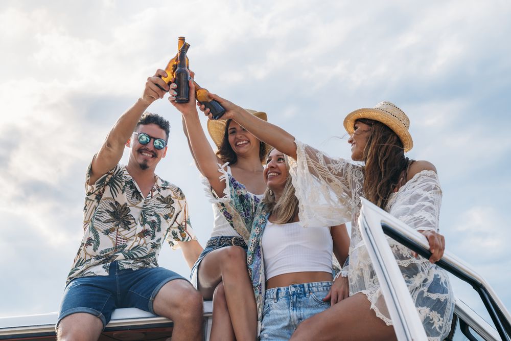 Smiling friends clinking bottles celebrating vacation on car roof in bright day