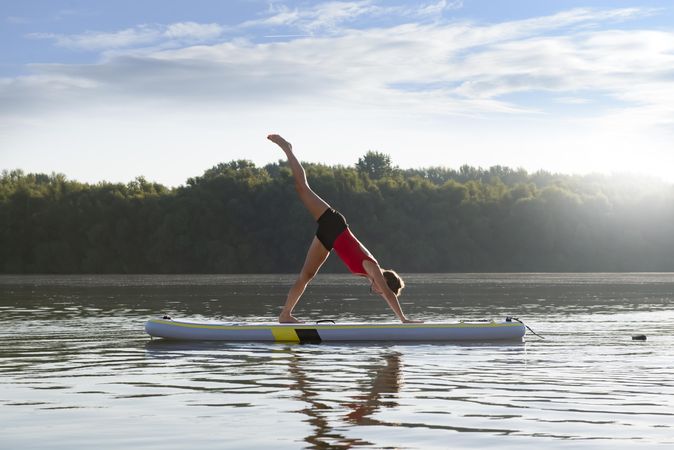 Woman stretching legs in yoga pose on paddleboard on fresh lake in the morning