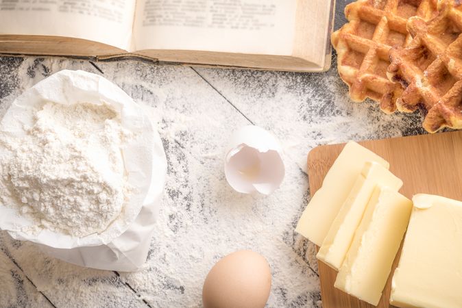 Top view of table with wheat flour, butter, eggs, a cookbook and freshly baked Belgian waffles