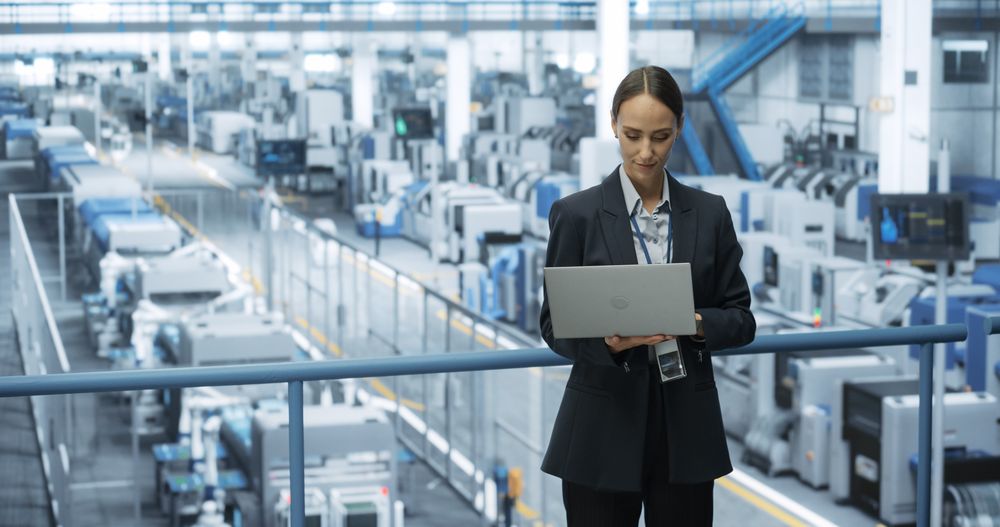 Portrait of a White Female Engineer Using Laptop Computer and Looking Around a Factory Facility with Robotic Arms Producing Modern Electronic Components for Tech Industries