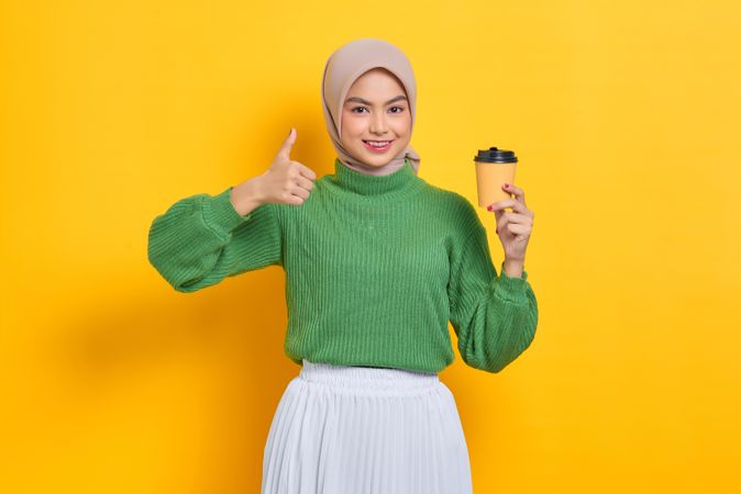 Woman in headscarf holding to go coffee cup and giving thumbs up sign