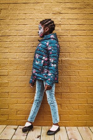 Side view of Black girl with face paint standing beside yellow brick wall