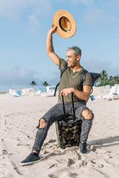 Portrait of cheerful young man waving hello seated on his luggage in south beach