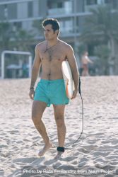 Male surfer walking on beach with board attached to his leg with leash 0VZZr0