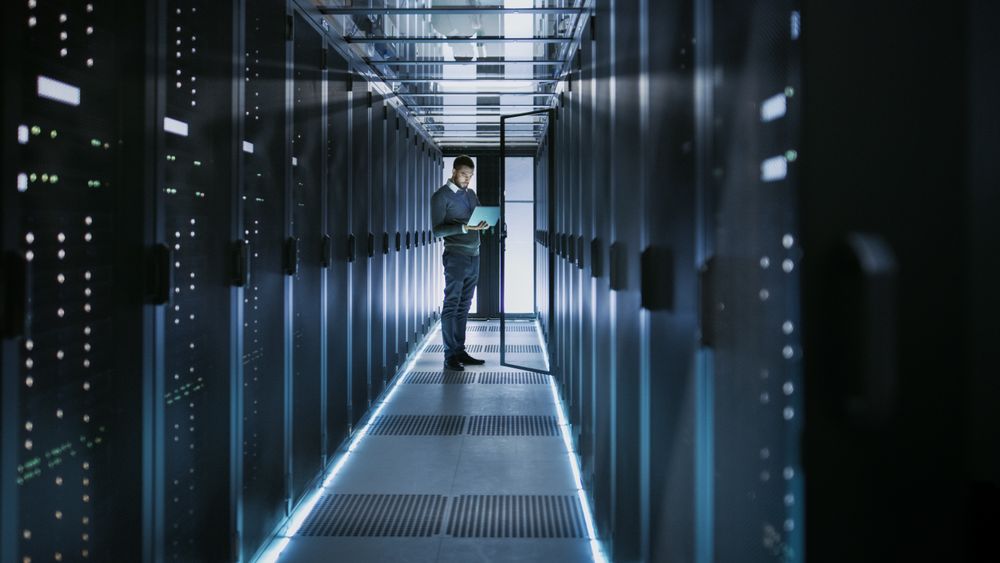 Long Shot of IT Technician Works on Laptop in Big Data Center full of Rack Servers.