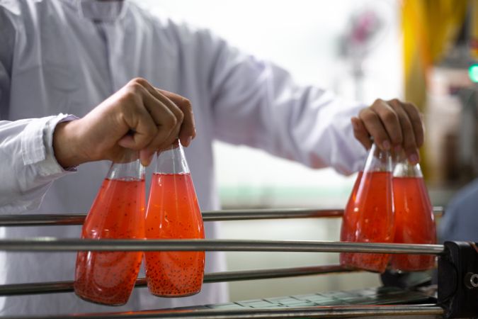 Hand of factory worker is picking up a bottle of fruit juice from a moving conveyor belt inside a beverage production facility.