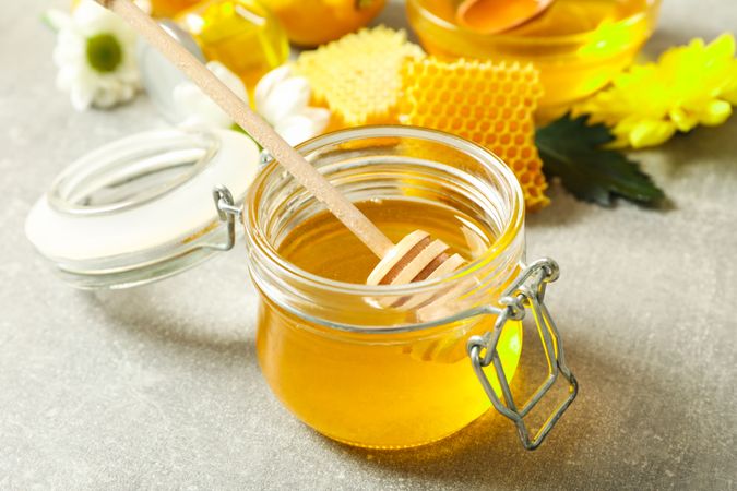 Glass jar with honey, dipper, honeycombs and flowers on grey background, closeup