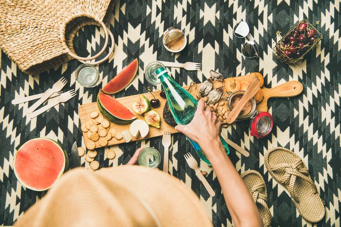 Woman pouring sparkling water at summer picnic on pueblo pattern blanket, with board of snacks