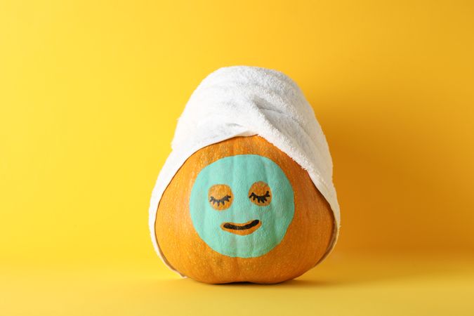 Pumpkin with facial mask and towel on yellow background, copy space