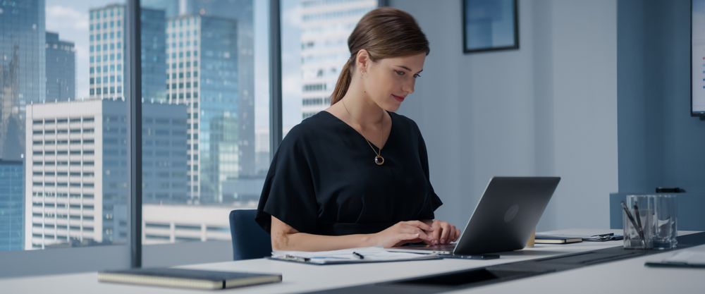 Businesswoman Standing In Her Corner Office Next To Big Window With City View And Using Laptop Computer.