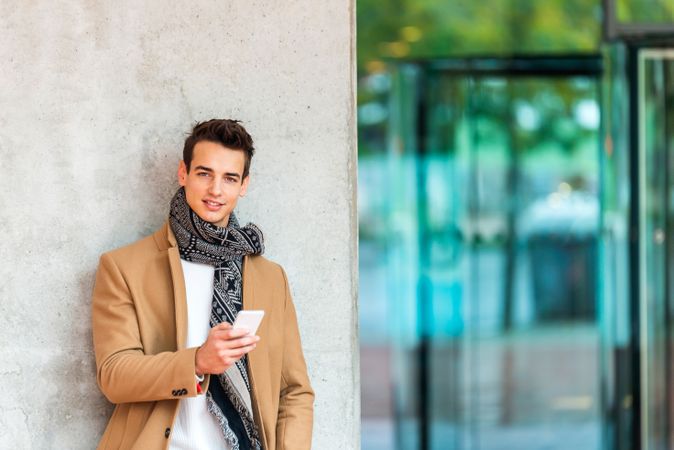 Stylish man leaning on wall outside texting and wearing a camel coat
