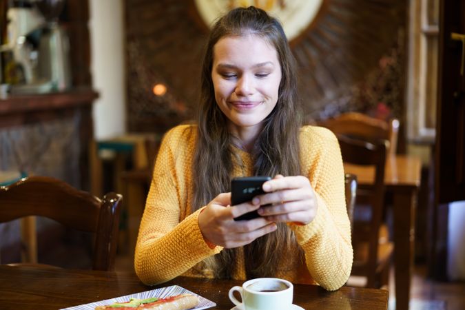 A young woman is enjoying a delicious cup of coffee and texting on her smartphone in a cozy cafe