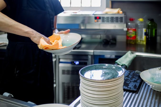 Crop woman in apron wiping plates in kitchen