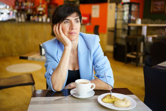 Female in trendy blue jacket sitting at table with cup of hot drink and slice of cake