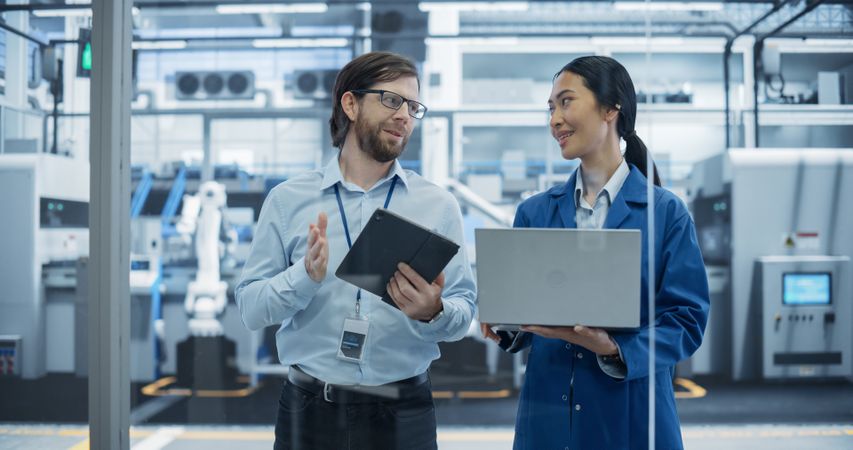 Engineer and Asian Female Project Manager Standing in Office, Talking and Using Computers and Monitoring Production at a Modern Automated Electronics Manufacture with AI Robotic Arms