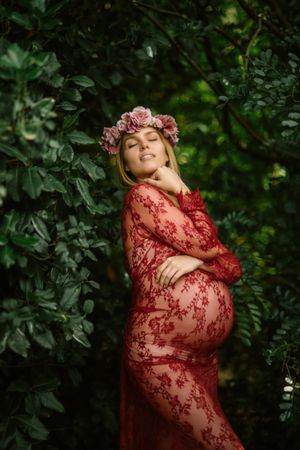 Pregnant woman with closed eyes touching belly and chin while standing in garden in sunny day