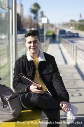 Young student sitting at the bus stop holding a notebook while looking at the camera with a smile 5Q28wn