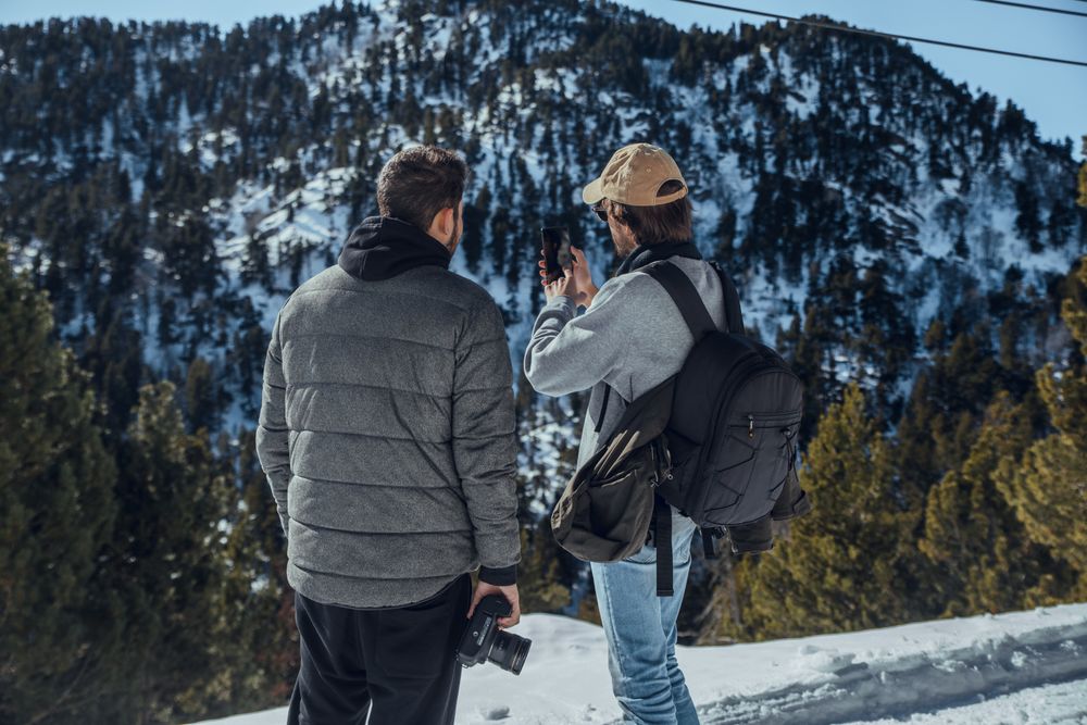 Men with smartphone and camera on hill between trees in winter