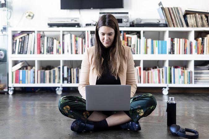 Portrait of a smiling woman sitting cross legged in front of book shelf working on laptop with water bottle