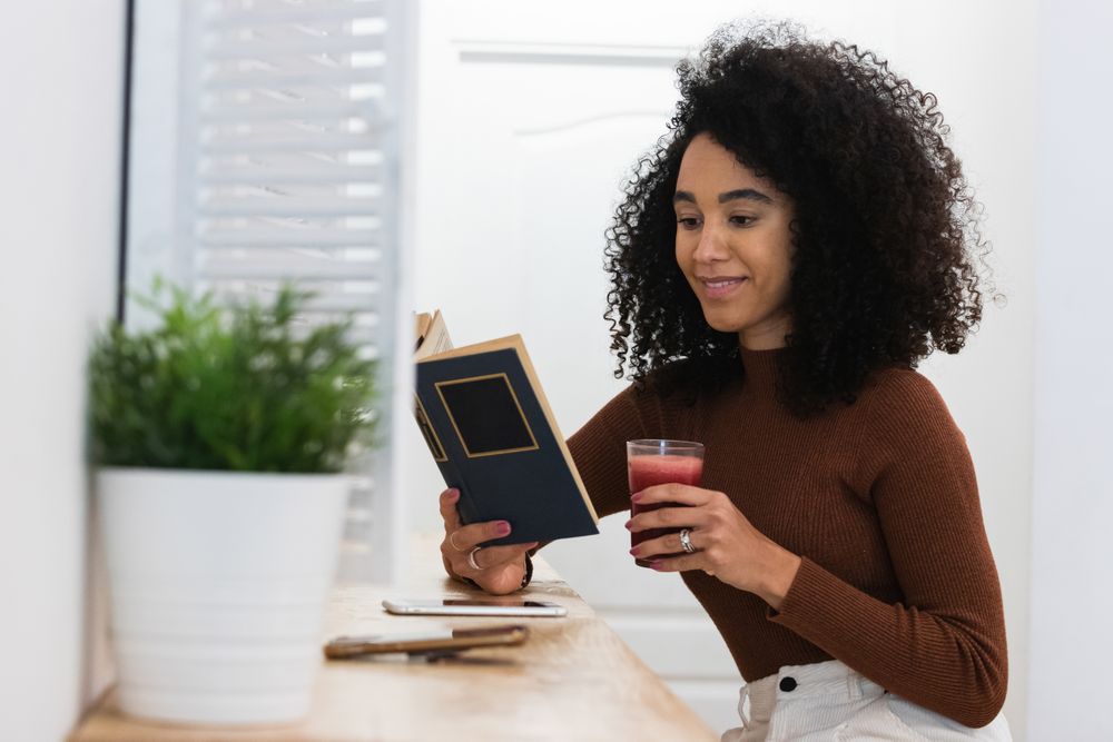 Black woman drinking smoothie and reading book in bar