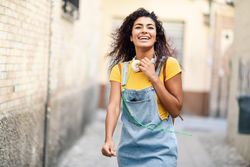 Smiling Arab woman with headphones and curly hairstyle walking outdoors in lane with graffiti