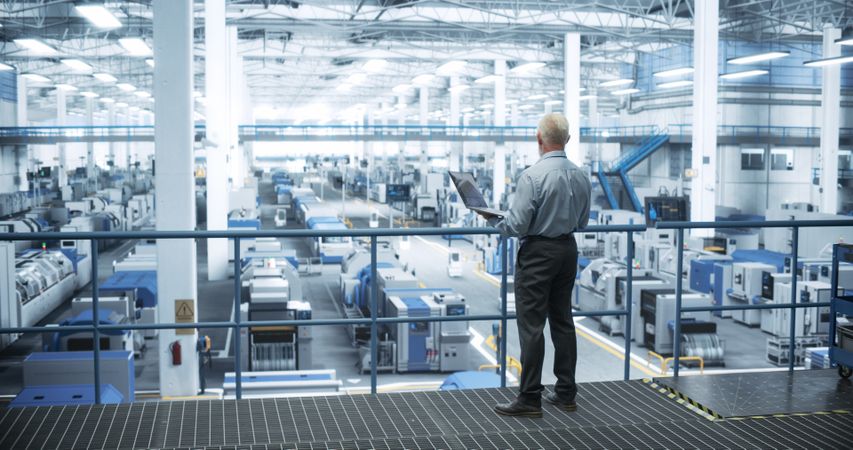 Experienced Male Engineer Standing on a Platform, Using Laptop Computer and Overlooking Production at a Modern Automated Electronics Manufacture with AI Support. Factory Executive at Work