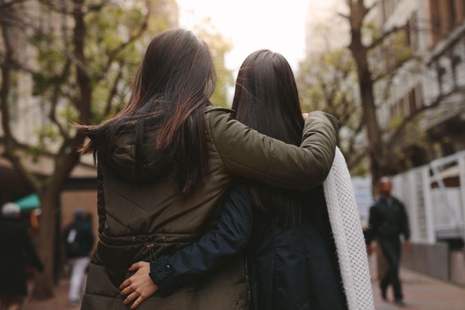 Backs of two dark-haired women hugging and walking down street