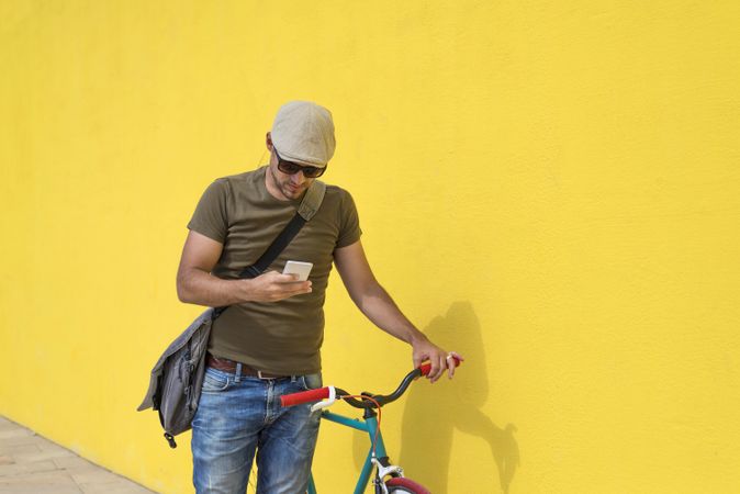 Man in hat and sunglasses checking phone standing with bike next to yellow wall