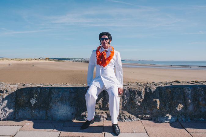 An Elvis impersonator in all white with orange lei sitting on rock wall by the beach